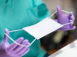 A person wearing purple gloves holds a white surgical mask.